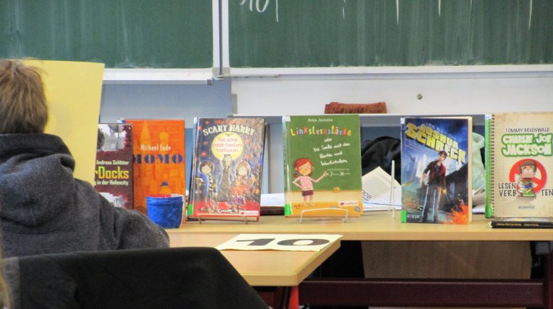 Foto eines Klassenraums in Blickrichtung zur Tafel. Bücher stehen mit dem Buchcover zum Betrachter hin aufgereiht auf einem Tisch, davor seitlich im Bild ein sitzender Schüler. 