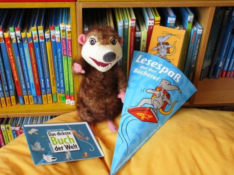 Foto Handpuppe Bücherfresser mit kleiner Papier-Schultüte, Gutschein für einen kostenlosen Büchereiausweis und kleinem Buch für Schulanfänger vor Kinderbuchregal. Die Schultüte trägt den Schriftzug: Lesespaß aus der Bücherei.