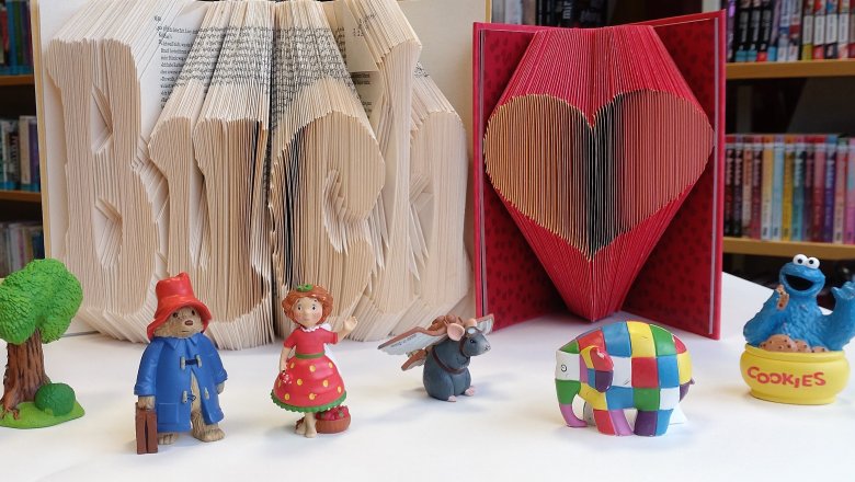 Foto als Symbolbild für die vielfältigen Angebote der Stadtbücherei: Auf einem Tisch stehen zwei Bücher, jeweils mit dem Buchschnitt zum Betrachter. Die Seiten sind so gefaltet, dass das Wort Buch zu lesen und ein Herz zu sehen sind. Diese Buchfaltkunst heißt Orimoto. Davor stehen sechs bunte Toniefiguren.