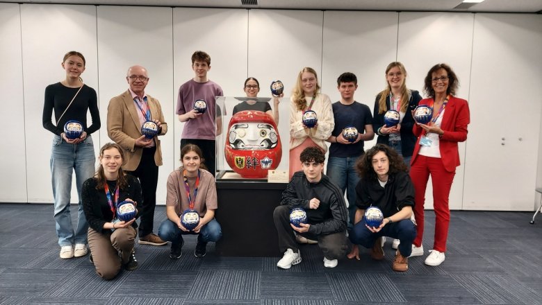 Daruma-Puppen sind die bekanntesten japanischen Glücksbringer. Die Jugenddelegation hat blauen Rohlinge mit dem Gesicht bemalt. Das Gruppenfoto zeigt außerdem den großen roten Daruma, der im Rahmen des 50-jährigen Städtepartnerschaftsjubiläums 2015 erstellt wurde. 